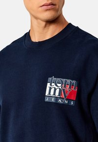 Felpa blu navy con colletto a girocollo, caratterizzata da un logo quadrato che include elementi rossi, bianchi e blu. Realizzata in morbido cotone, ha una vestibilità informale.