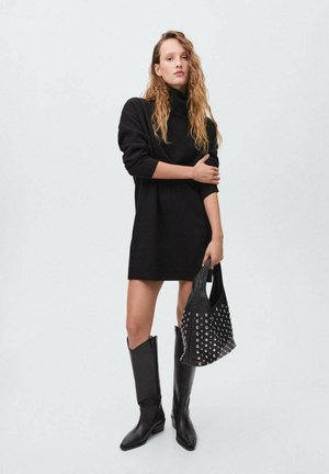 Vestido suelto negro de punto con cuello alto, combinado con botas de cuero negras hasta la rodilla y un bolso negro con tachuelas. Textura suave, diseño moderno.