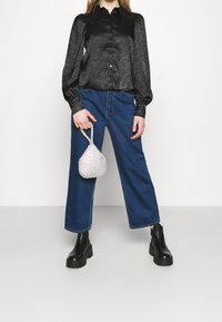 Musta satiinipusero, jossa on napitus ja hienovarainen kuvio, yhdistetty korkeavyötäröisiin sinisiin leveälahkeisiin farkkuihin. Kädessä hopeinen strassikoristeinen laukku.