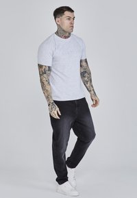 T-shirt grigio a maniche corte con logo in rilievo, abbinato a jeans neri a sigaretta e scarpe da ginnastica bianche. Presenta tatuaggi su braccia e collo.