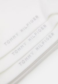 Weiße Baumwollsocken mit geripptem Muster, die wiederholt das Branding "TOMMY HILFIGER" in hellgrau zeigen. Drei Paar gestapelt.