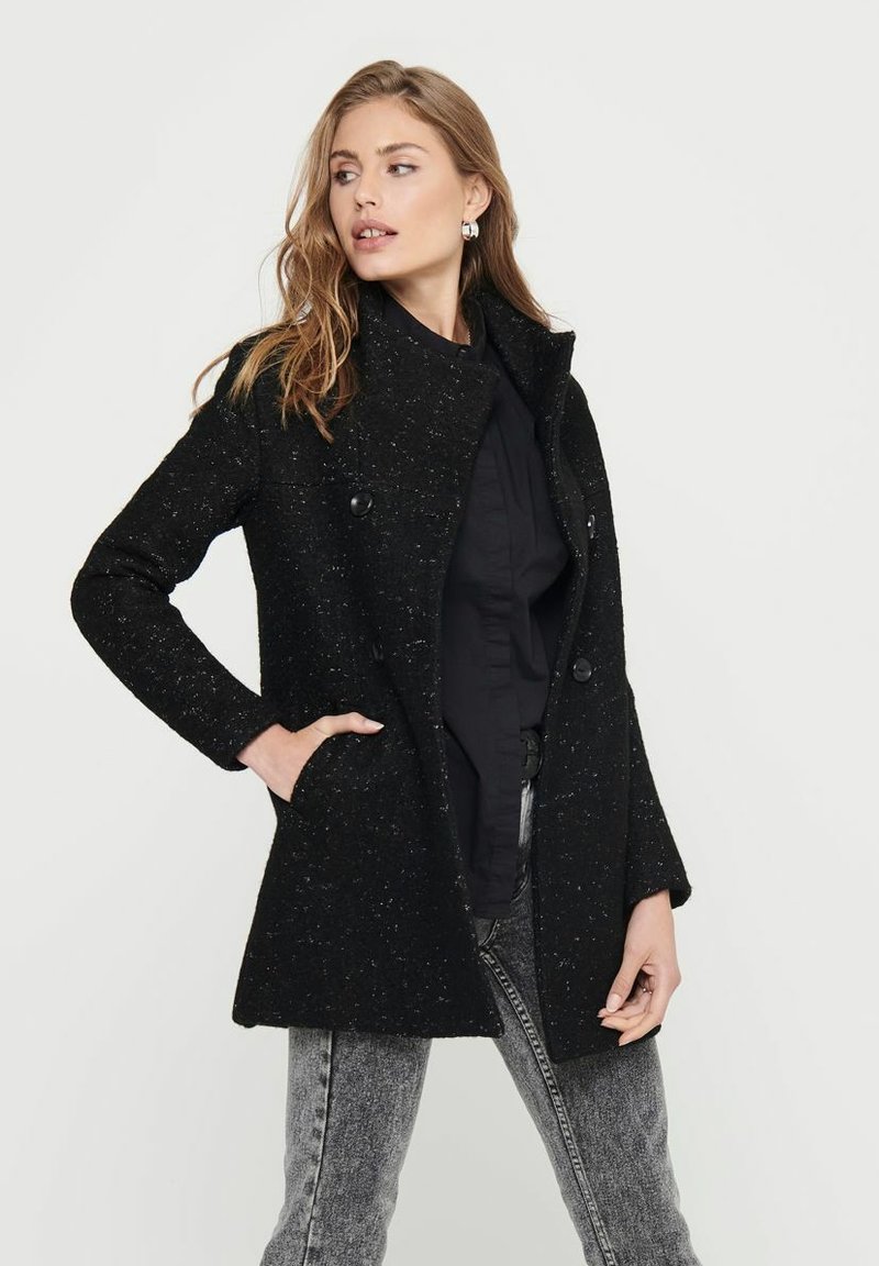 Manteau noir en mélange de laine avec une finition texturée, un col structuré, deux boutons et des poches latérales ; associé à un haut noir et un jean gris.