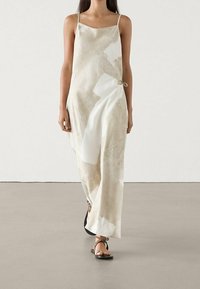 Robe nuisette en satin beige avec des motifs abstraits blancs, dotée de fines bretelles, d'un détail de nœud sur le côté et d'une coupe décontractée. Des sandales plates noires complètent le look.