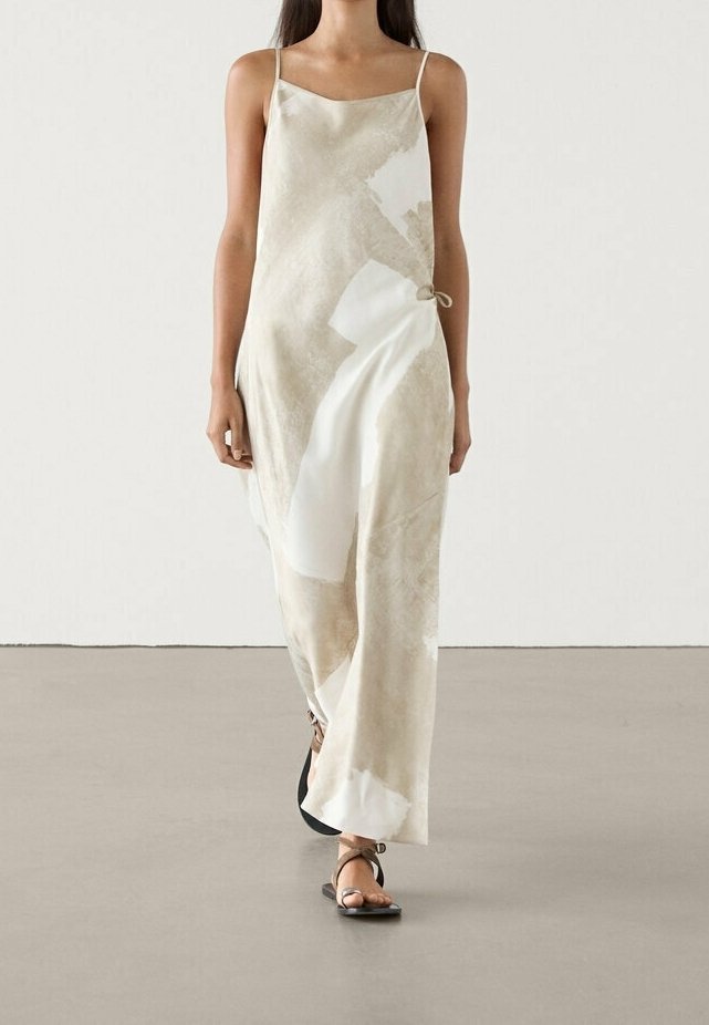 Robe nuisette en satin beige avec des motifs abstraits blancs, dotée de fines bretelles, d'un détail de nœud sur le côté et d'une coupe décontractée. Des sandales plates noires complètent le look.