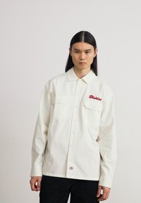 White button-up shirt with two front pockets, featuring red embroidered text. Smooth fabric with a relaxed fit and pointed collar.