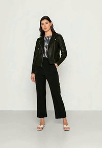 Jaqueta de moto em pele preta com zíperes prateados, usada sobre uma blusa cinza com efeito tie-dye. Calças largas pretas e sandálias brancas completam o visual.