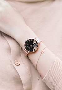 Runde Uhr mit schwarzem Blumen-Zifferblatt, roségoldenem Gehäuse und Mesh-Armband. Begleitet von einem zarten Armband mit einem kleinen runden Anhänger.