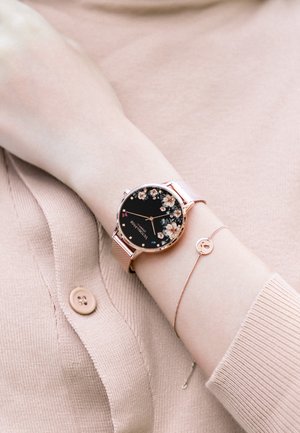Montre ronde avec un cadran floral noir, un boîtier en rose gold et un bracelet en maille. Accompagnée d'un bracelet délicat avec un petit charm circulaire.
