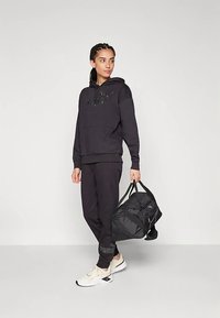 Melna kapuce ar priekšējo kabatu un logotipu, kombinēta ar atbilstošām sporta biksēm un bēšām kedām. Modelis tur melnu sporta somu.