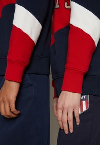 Due persone indossano felpe colorate, con righe rosse, bianche e blu navy. Entrambe hanno polsini a costine e pantaloni blu navy con un logo.