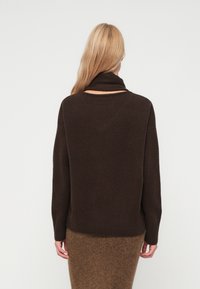 Suéter castanho-escuro com colarinho alto e dobrado, e bainha canelada, apresenta um corte descontraído e uma textura suave. Modelo usa uma saia castanha que complementa.