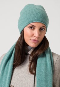 Berretto a maglia verde menta con una texture morbida, abbinato a una sciarpa coordinata. L'outfit si completa con un cappotto grigio chiaro e un dolcevita.