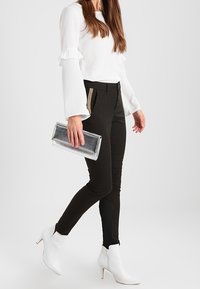Svarta tight passande byxor med sidogylf, i kombination med en vit blus med långa ärmar och volangärmar, samt en genomskinlig clutch. Vita ankelstövlar.