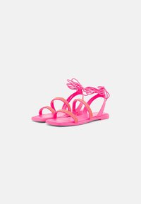 Sandales roses avec des brides courbées, une semelle plate et des accents décoratifs. Conçues avec un lien en lacet autour de la cheville pour un maintien sécurisé.