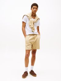 Beige Baumwollshorts mit elastischem Bund, kombiniert mit einem weißen V-Ausschnitt-T-Shirt und einem beige Sweatshirt, das über die Schultern gelegt ist. Braune Sneakers.
