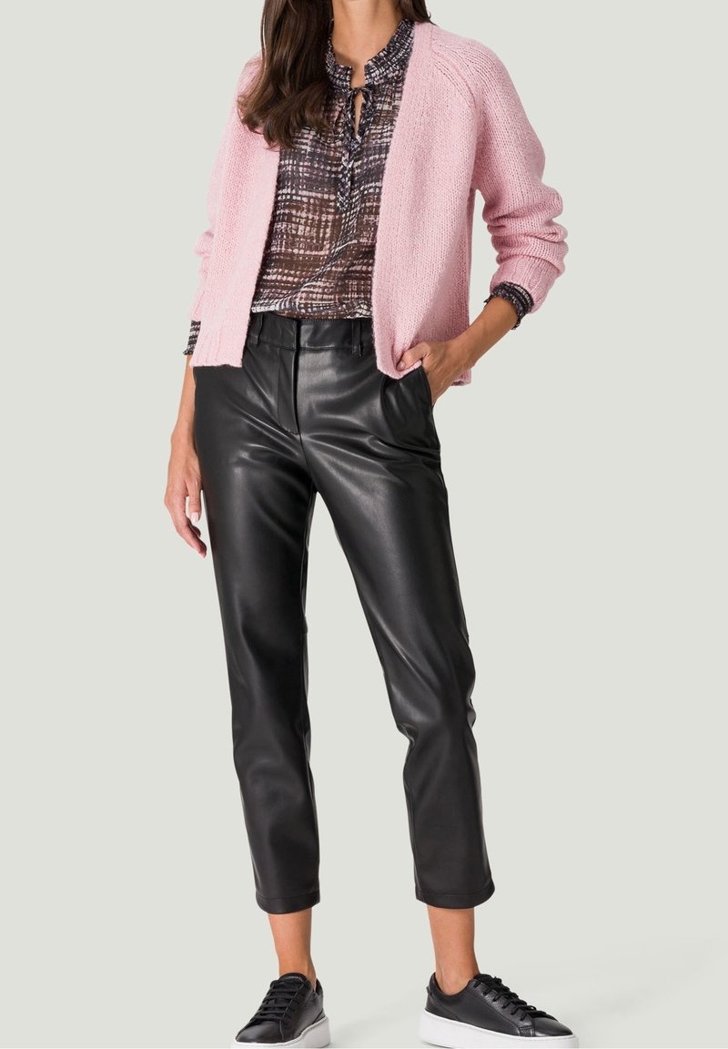 Femme portant un cardigan rose en maille sur un chemisier à motifs, un pantalon en cuir noir et des baskets noires à semelles blanches.