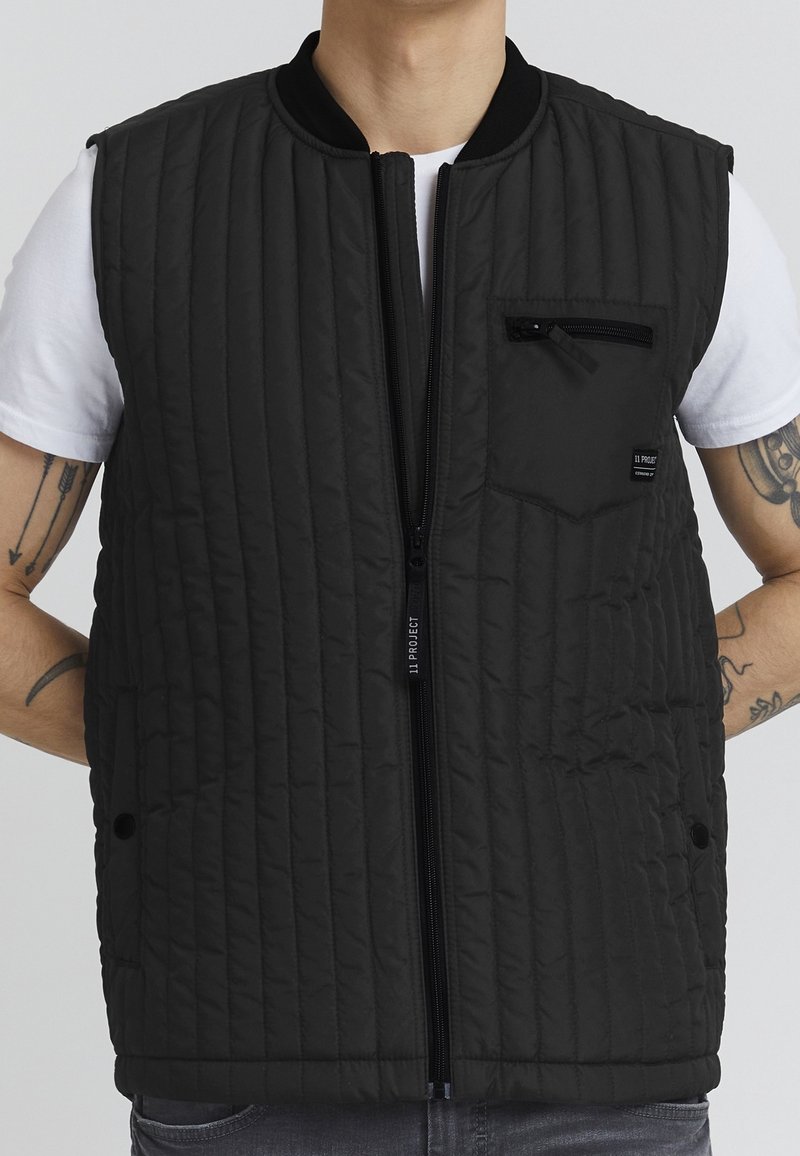 Homme portant un gilet noir matelassé sans manches avec fermeture éclair à l'avant et poche poitrine zippée par-dessus une chemise blanche à manches courtes.
