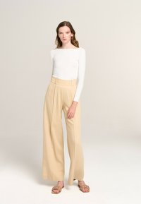 Beige wide-leg trousers with pleats at the front, paired with a fitted white long-sleeve top and tan flat sandals, standing on a neutral background.
