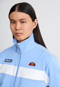 Light blue zip-up jacket with a high collar, white stripes, and embroidered red and orange logo. Smooth texture with a sporty design.