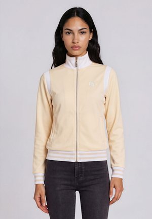 Femme aux longs cheveux foncés portant une veste beige clair zippée avec des accents blancs et un pantalon sombre, debout devant un fond uni.