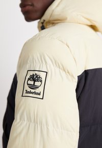 Bunda s vypchávanou štruktúrou v krémovej farbe s kontrastnou čiernou časťou, prešívaná a s nášivkou Timberland na rukáve.