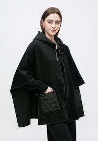 Must kapuutsiga mantel, millel on tikitud mustad taskud, nöörkinnitusega ja soonikulised varrukaservad. Pehme materjal ja lahtine, voolav disain.