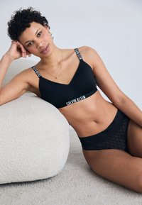 Svart Calvin Klein bralette med v-hals og logo-stropper, kombinert med svarte blondeundertøy med sirkulært mønster; myke teksturer.