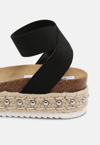 Sandale à bride élastique noire avec une semelle en liège et une plateforme en jute tissé texturé, décorée de perles argentées le long du bord.