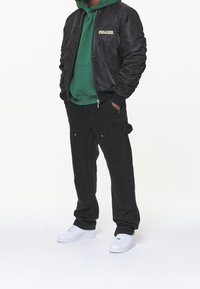 Giubbotto bomber nero con ricamo "PEGADOR", felpa verde con cappuccio e pantaloni cargo neri con tasche. Scarpe da ginnastica bianche completano l'outfit.
