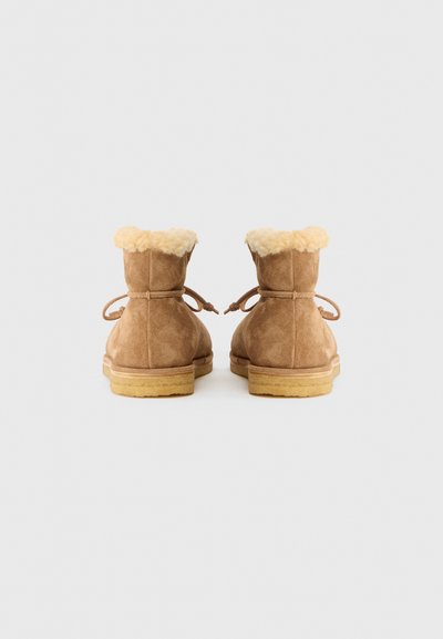 Botas de tornozelo em camurça castanha com acabamento em pêlo sintético creme, solas de borracha planas e atacadores, mostradas de costas sobre um fundo branco.