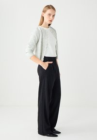 Une jeune femme se tient avec les mains dans les poches, portant une chemise blanche, un cardigan gris clair, un pantalon large noir et des chaussures noires à bout pointu.