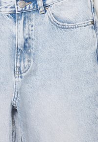 Svetlo modri jeans iz jeansa od blizu, prikazujoč sprednji gumb, zanke za pas, sprednji žep in podrobno šivanje.