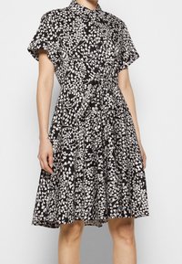 Frau in einem schwarzen Kleid mit weißen, spiralförmigen herzförmigen Mustern, kurzen Ärmeln, einem Kragen und einem gebundenen Knoten an der Taille.