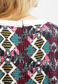 Tissu à motifs multicolores présentant des formes géométriques en rouge, sarcelle, noir et or, avec un col texturé blanc et un détail de fermeture éclair au dos.