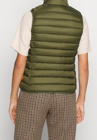 Zielona kamizelka puchowa z poziomym pikowaniem i wysokim kołnierzem. Podkreślona pomarańczowym akcentem. Noszona na jasnoszarym pewnohem i wzorzystych spodniach.