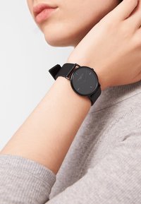 Orologio analogico nero con un quadrante rotondo minimalista, finitura opaca e cinturino in pelle nera. Indicatori delle ore visibili e lancette sottili.