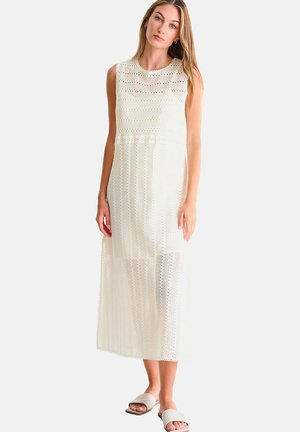 Jeune femme portant une robe midi blanche en crochet sans manches et des sandales beiges à enfiler, debout contre un fond clair uni.