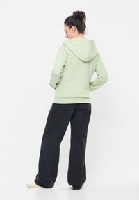 Felpa verde chiaro con cappuccio e tasca frontale a marsupio, abbinata a jeans neri larghi. Il tessuto sembra morbido con una consistenza liscia.