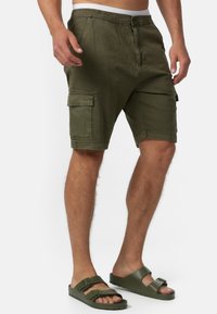 Hombre vistiendo pantalones cortos tipo cargo verde oliva y sandalias de hebilla a juego en verde oliva, de pie contra un fondo blanco liso.