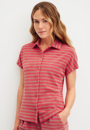 Frau mit welligem blondem Haar, die ein rot-beige gestreiftes kurzärmliges Hemd mit Knöpfen und passende Hosen trägt, steht mit einer Hand in der Tasche.