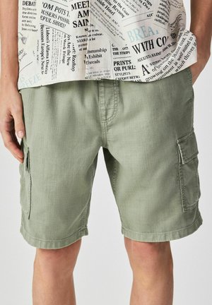 Mann trägt hellgrüne Cargoshorts und ein weißes Hemd mit schwarzem Zeitungsprint, steht mit entspannten Händen an den Seiten.