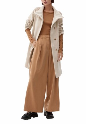 Abrigo de lana beige con solapas grandes y botones, combinado con una camisa marrón de manga larga y pantalones anchos color tostado. Zapatos negros completan el conjunto.