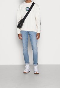 Felpa bianca a girocollo con ricamo di un globo multicolore, jeans slim-fit azzurri, e sneakers bianche con suola marrone, borsa a tracolla.