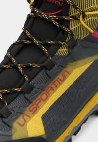 Shoe de caminhadas preto e cinza com um cabedal em malha e detalhes em amarelo, sobreposições texturizadas e atacadores vermelhos. A marca está visível na lateral.