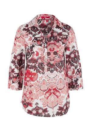 Femme portant une chemise à fleurs rouge et blanche boutonnée, un pantalon blanc taille haute retroussé et des baskets blanches, debout les mains dans les poches.