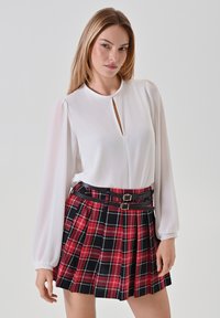 Weiße Bluse mit rundem Halsausschnitt und Schlüssel-Ausschnitt, kombiniert mit einem plissierten roten Karorock mit schwarz-weißen Streifen und zwei schwarzen Gürteln.