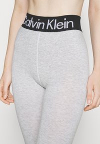 Šedé legíny vyrobené z měkkého, elastického materiálu s černým elastickým pasem, na kterém je výrazně zobrazeno bílé logo "Calvin Klein".