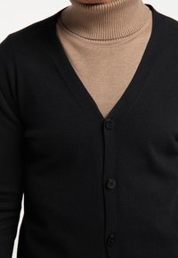 Cardigan nero a collo a V con cinque bottoni, indossato sopra un dolcevita beige. Presenta una texture a coste e un tessuto morbido e lavorato a maglia.