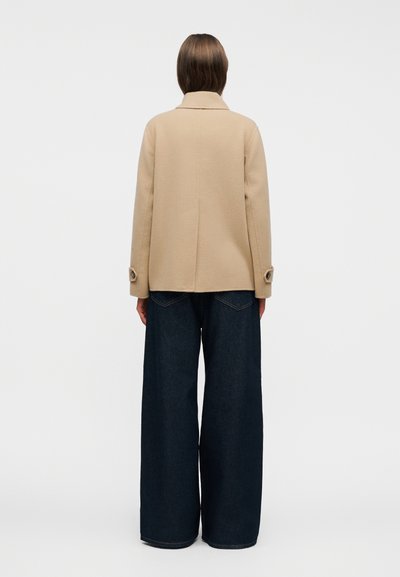 Άτομο που φοράει μπεζ μπουφάν με μανσέτες με κουμπιά και φαρδιές σκούρες μπλε τζιν, στέκεται με την πλάτη προς τα πίσω σε λείο λευκό φόντο.