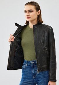 Giubbotto di pelle nera con colletto rotondo, chiusura con zip e tasche laterali; indossato sopra un top verde e jeans a vita alta blu.